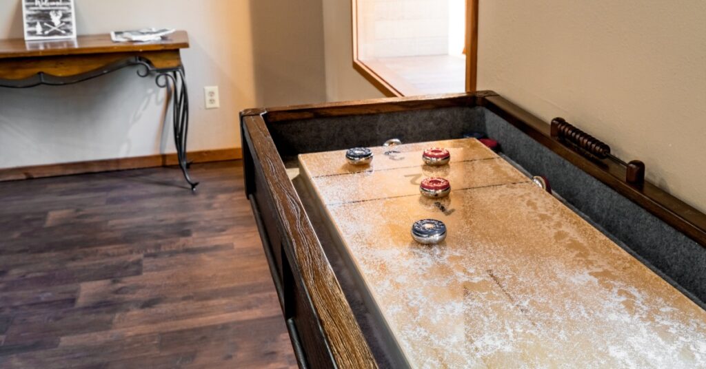 Large wooden shuffleboard table with red and blue pucks inside the game room of a house with beige walls and wooden floors.