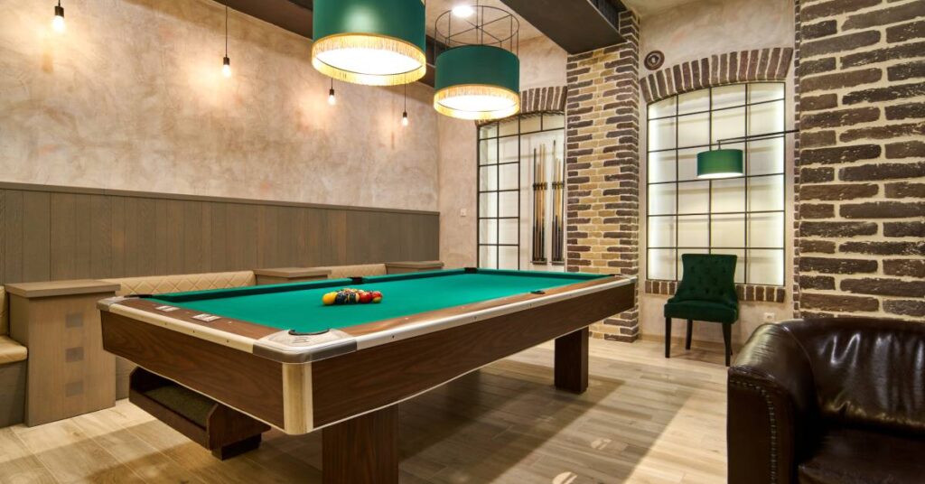 New pool table with the balls on top of it inside a cozy room with exposed brick, cushioned seats, and warm overhead light.