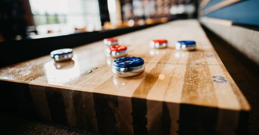 Close-up to the edge of a shuffleboard with three blue pucks and three red ones inside a room with a blurry background.