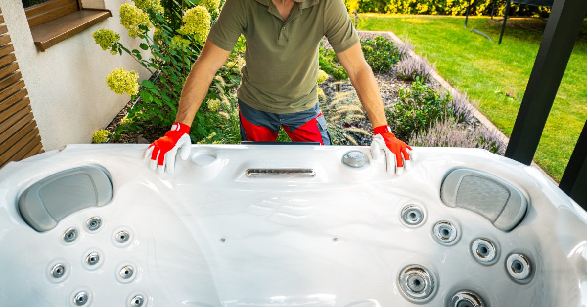 Man wearing a red cap and red safety gloves standing behind an empty hot tub placed in the backyard of a home.